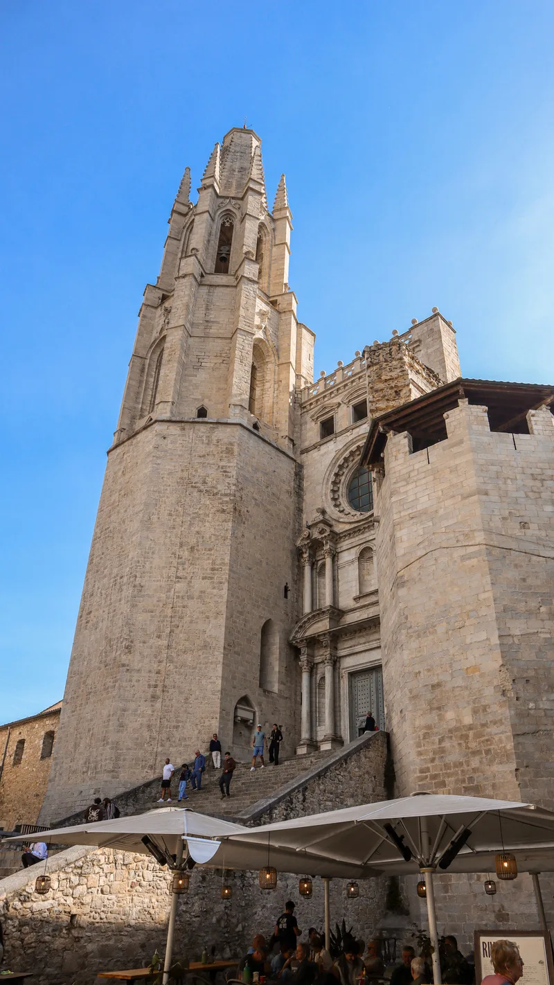 The Gothic bell tower of the Basilica of Sant Feliu above a stone staircase and café umbrellas