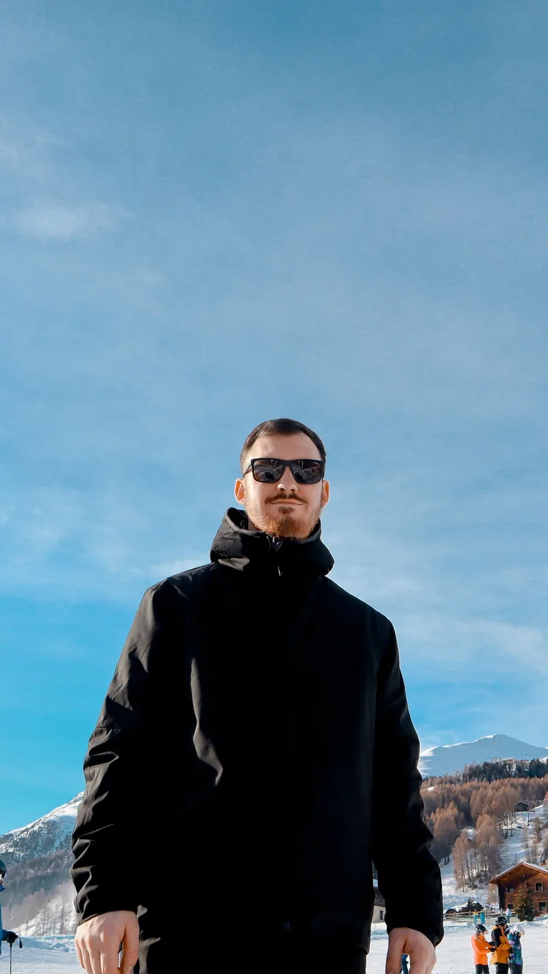 Bryce de Bruyn in sunglasses and a black jacket, standing in the snow with mountains behind