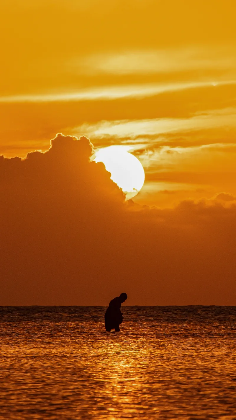 A lone figure standing in the sea at sunset under orange clouds