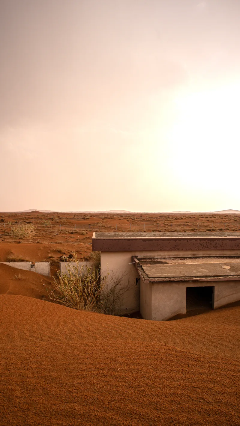 An abandoned house half-buried by red sand dunes under a hazy sun