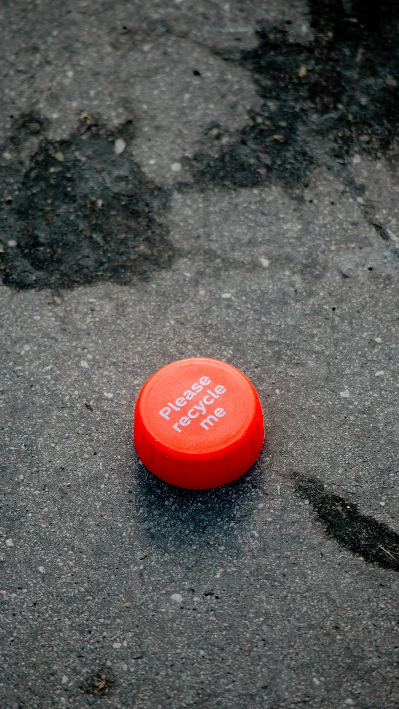 A red bottle cap reading 'please recycle me' on wet tarmac